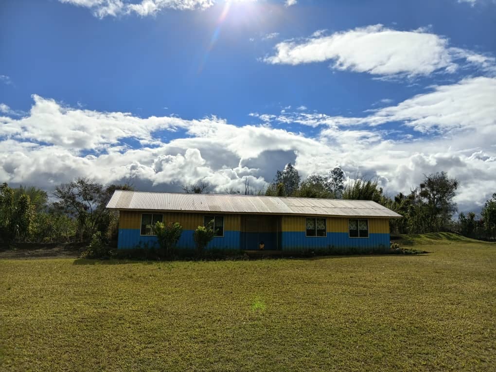 A single yellow and blue painted school building with a metal roof on open grassland under a bright sun and scattered clouds in Akaka