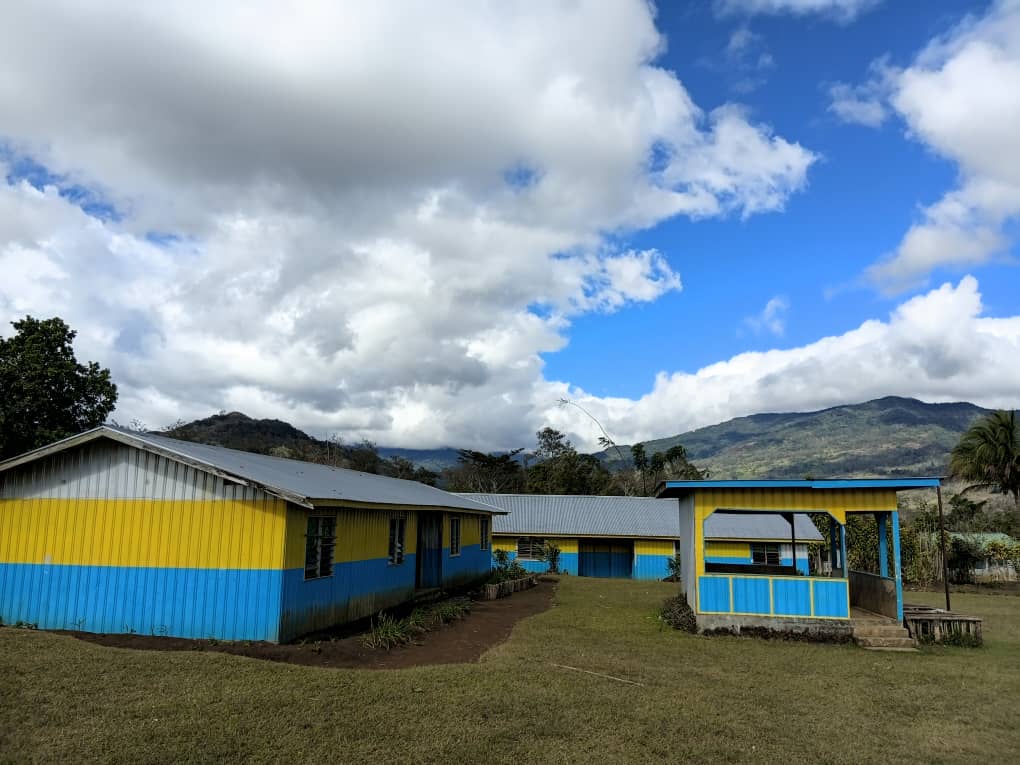 Yellow and blue painted school buildings with a covered shelter and mountain backdrop under a partly cloudy sky in Akaka