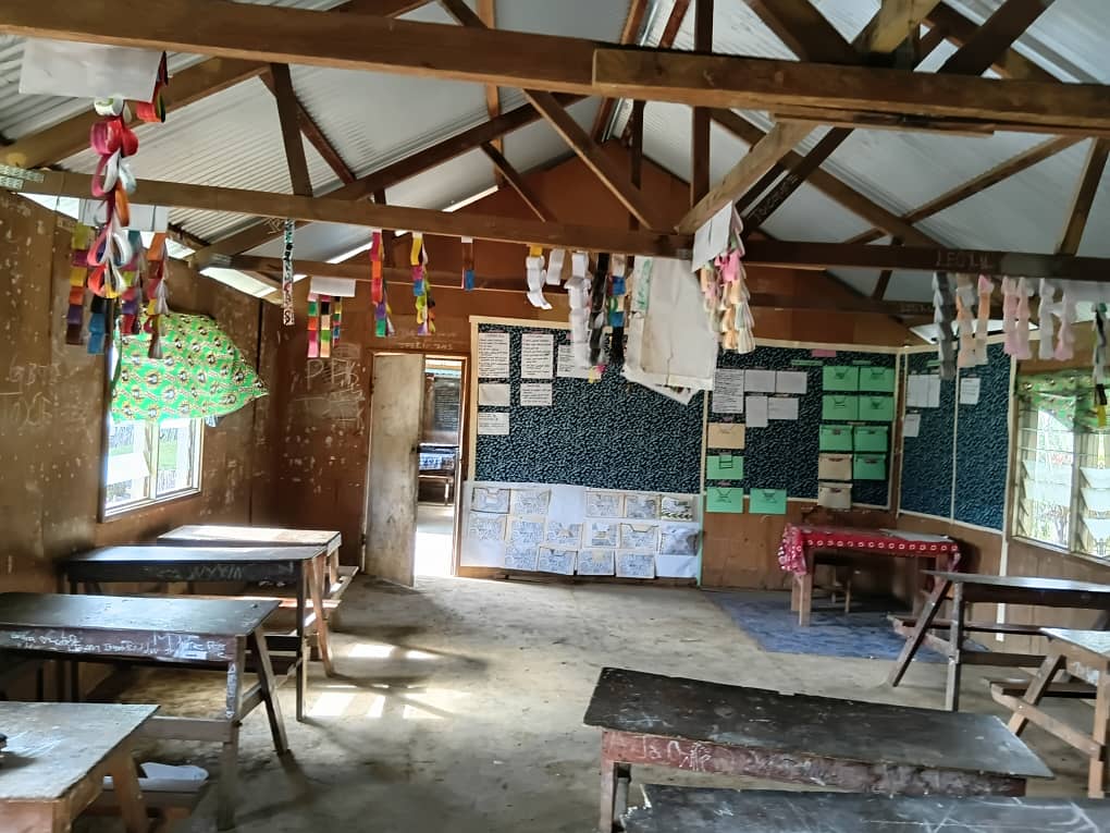 Interior of a classroom at Akaka Community School with wooden desks, a blackboard covered in papers, and colourful decorations hanging from the ceiling beams.