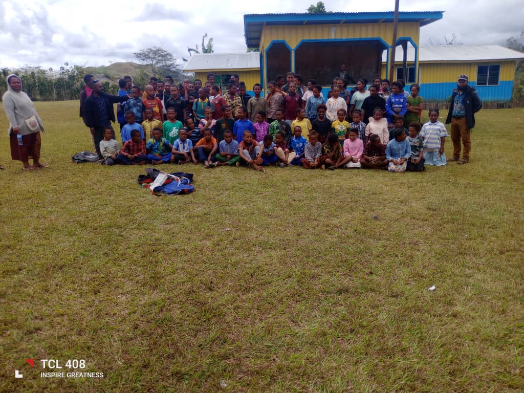 A large group of students and teachers posing together on the grass in front of a yellow and blue school building at Akaka Community School.