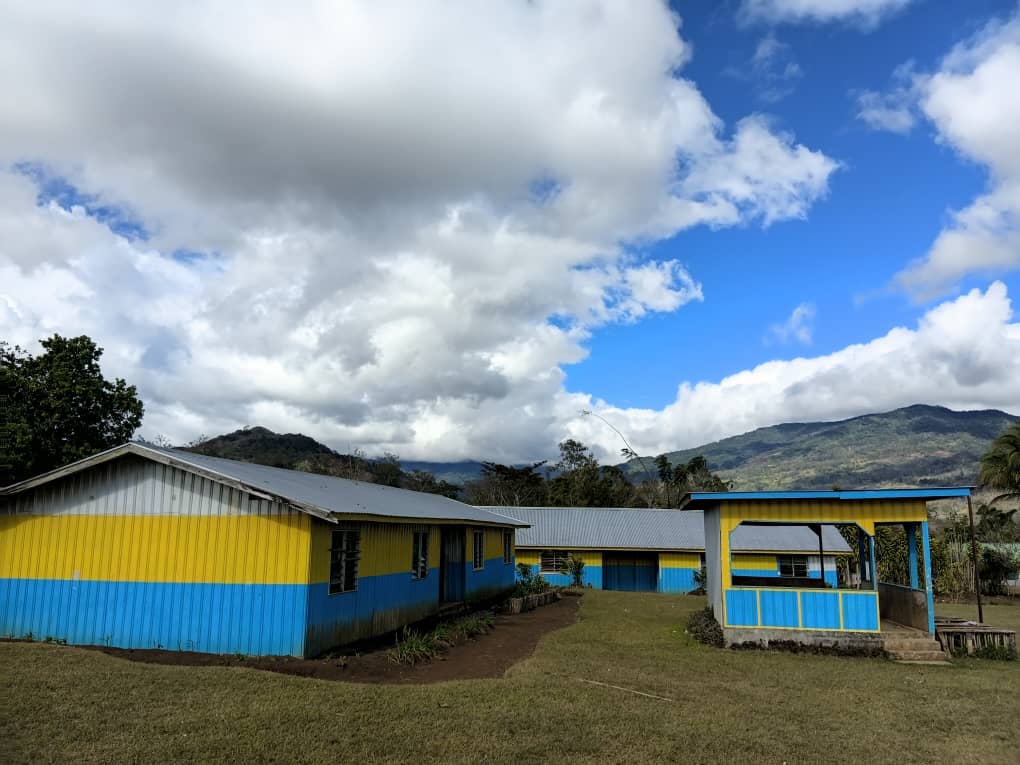 Yellow and blue painted school buildings with a covered walkway and green mountains under a partly cloudy sky in Akaka