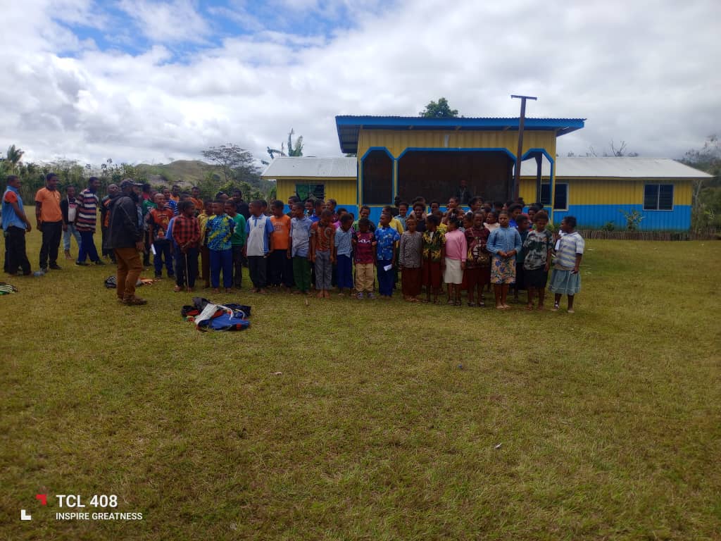 Students and teachers gathered on the grass in front of a yellow and blue school building at Akaka Community School.