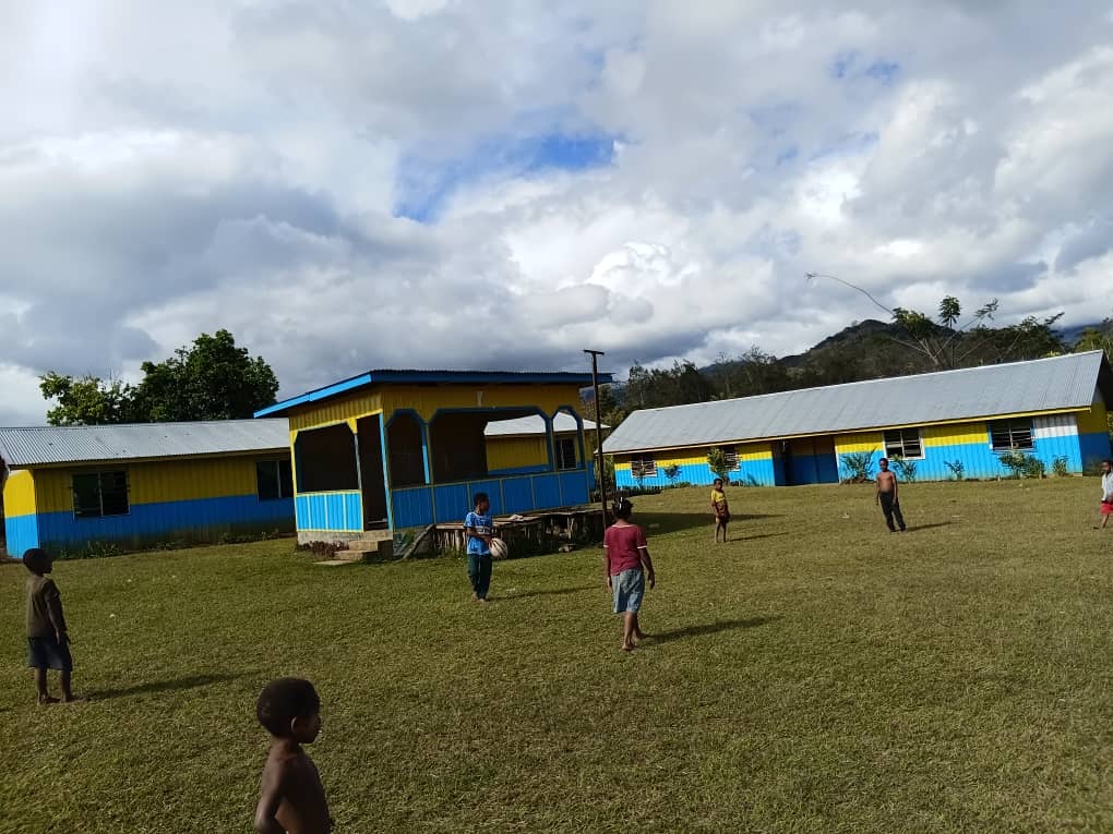 Children on a grassy field in front of yellow and blue painted school buildings under a cloudy sky in Akaka
